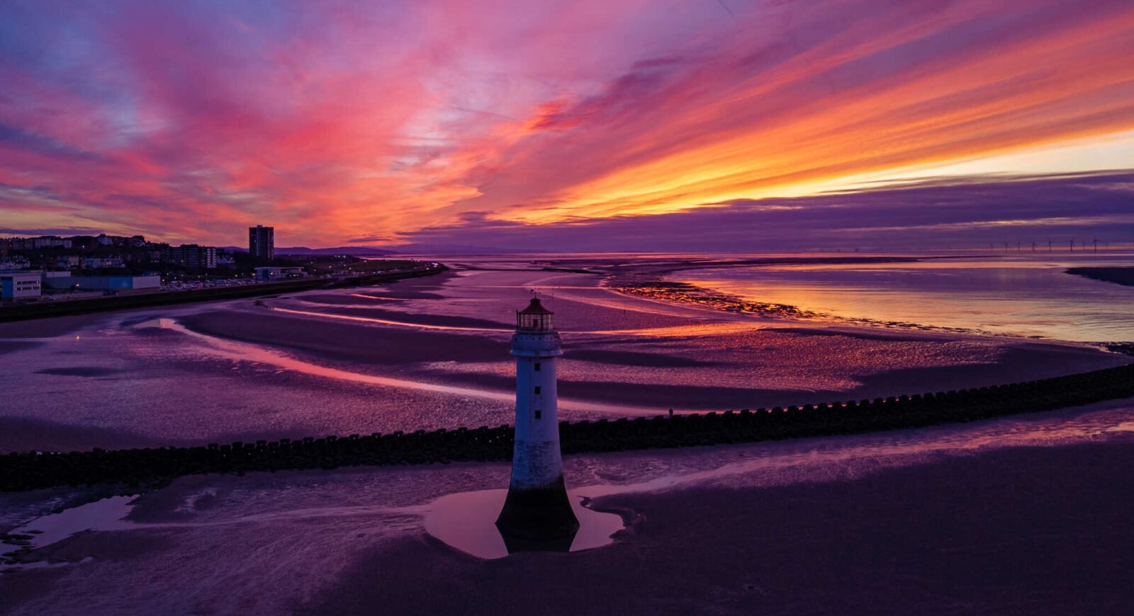 New Brighton Lighthouse at sunset with vivid purple, orange, and pink skies reflecting across the wet sands and tidal channels of the River Mersey coastline.