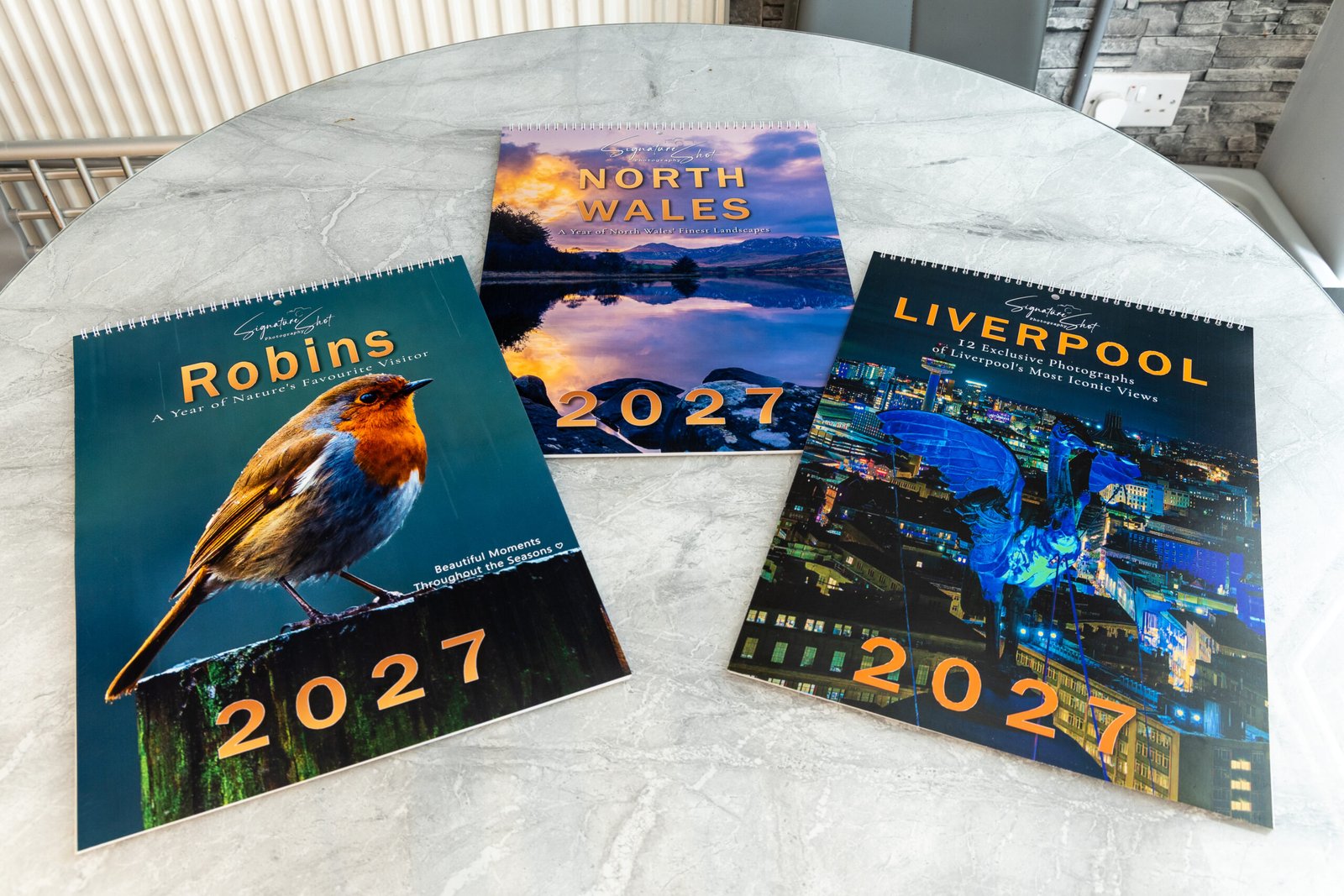 The Robin, North Wales and Liverpool 2027 calendar front covers photographed together on a marble table